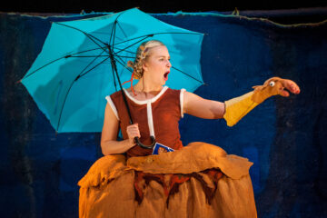 Keltahameinen näyttelijä pitelee sinistä sateenvarjoa ja näyttelee keltaisen käsinuken kanssa.