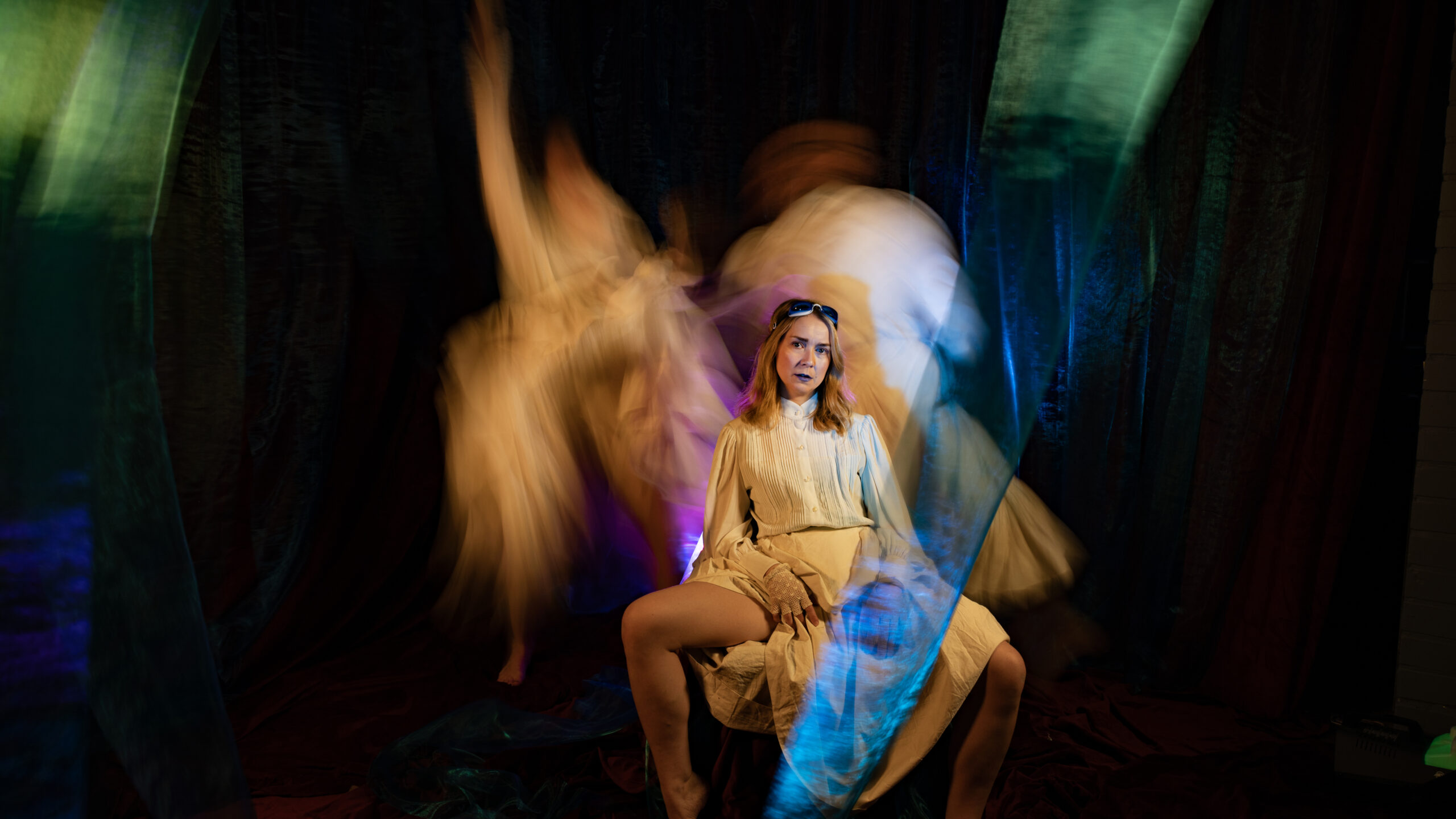 Vaalea nainen valkoisessa hameessa ja uimalasit otsalla istuu hajareisin. Taka-alalla kaksi valkoisiin pukeutunutta epätarkkaa hahmoa.