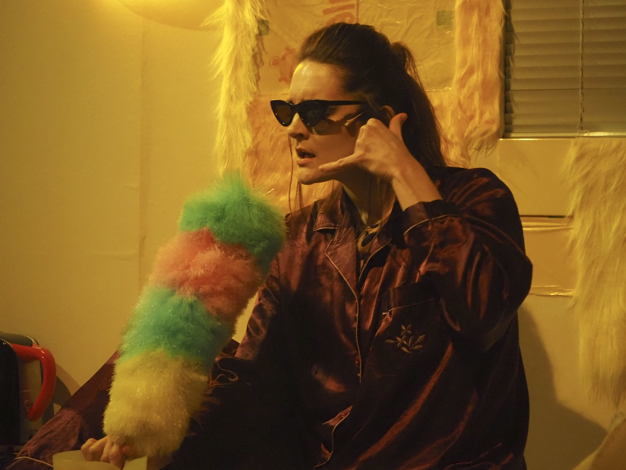 Ihminen, jolla on aurinkolasit päässä, esittää puhuvansa puhelimen luuriin, jonka on tehnyt kädestään ja leikkii pölyhuiskan olevan mikrofoni.